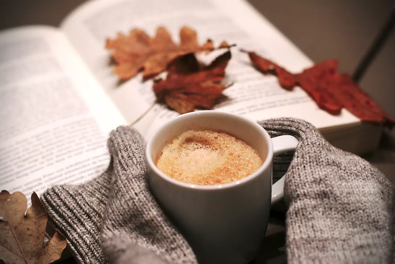 A cup of coffee sits between sleeves of a sweater with a book and fall leaves in a relaxing background.